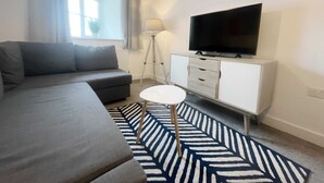 Un salon moderne avec un canapé, un téléviseur posé sur un meuble blanc, une lampe et un tapis à motifs.