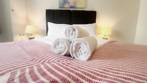 Une chambre d’hôtel avec un lit, des oreillers, des serviettes roulées, une tête de lit, deux lampes de chevet et un tableau au mur.