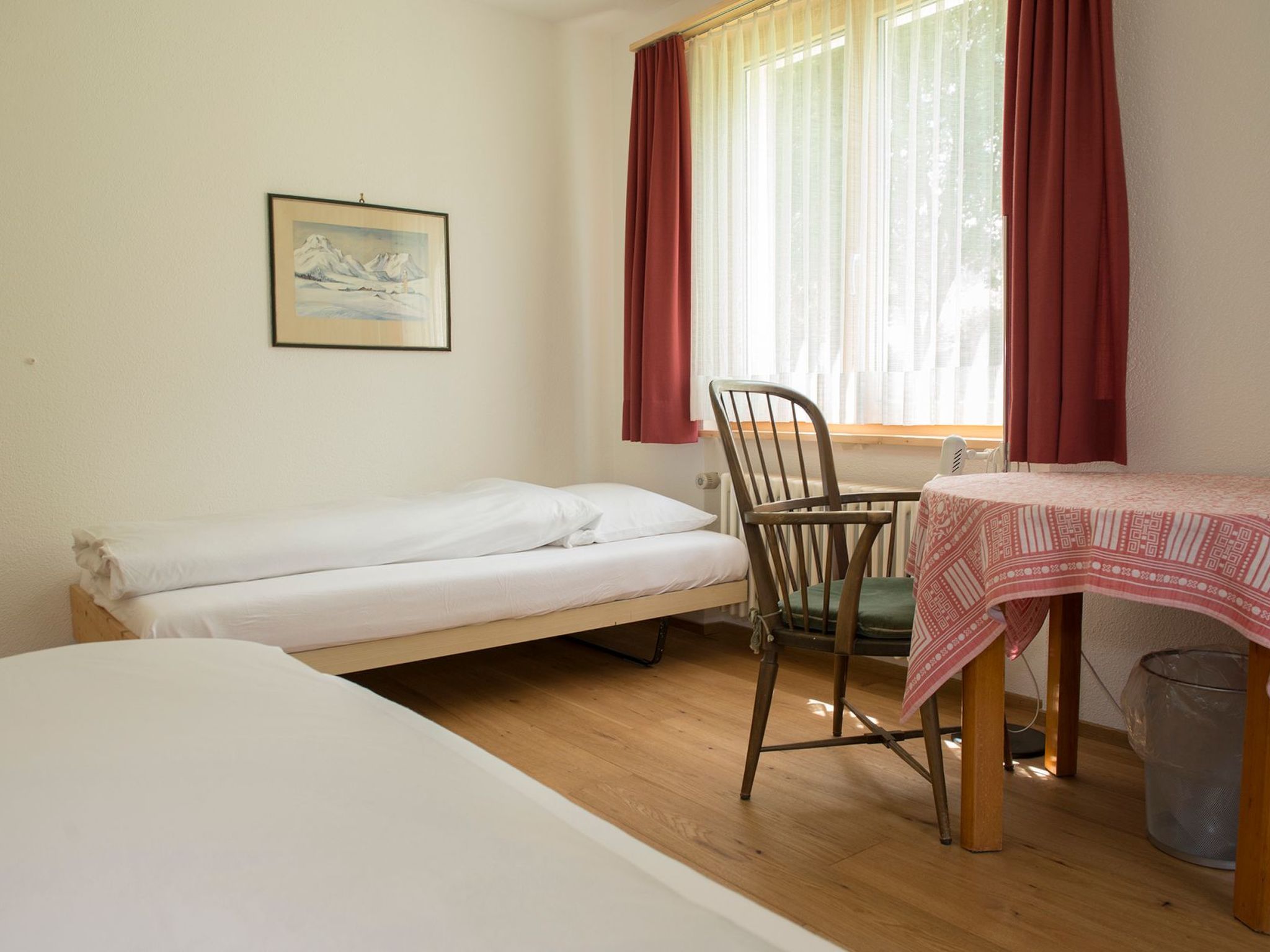A single-bed room with a wooden floor, a chair, a table with a patterned tablecloth, a radiator, and a window with red curtains.