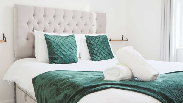 Un lit bien fait, avec une tête de lit capitonnée, deux coussins verts et un plaid vert.