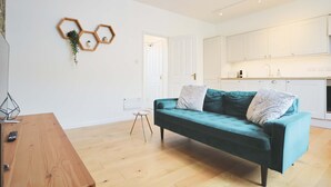 Un salon moderne avec un canapé de couleur turquoise, une table basse en bois et un petit tabouret. On aperçoit une cuisine à l’arrière-plan, avec des meubles blancs et un plan de travail.