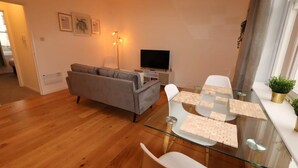 Een moderne woonkamer met een bank, een glazen eettafel met stoelen, een flatscreen televisie en ingekaderde foto's aan de muur.