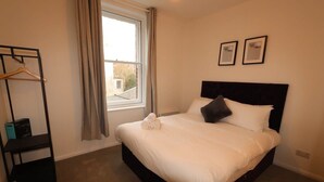Een hotelkamer met een bed, een raam, een kapstok en twee foto's in lijsten aan de muur.