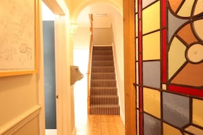Uma escadaria que leva a um andar superior, uma porta com desenho em vitral e uma parede com um mapa ou diagrama.
