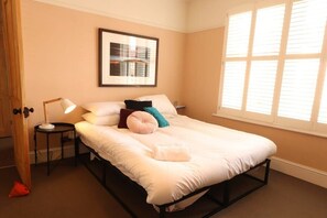 Quarto com cama, mesa de cabeceira, luminária, quadro na parede e janela com persianas.