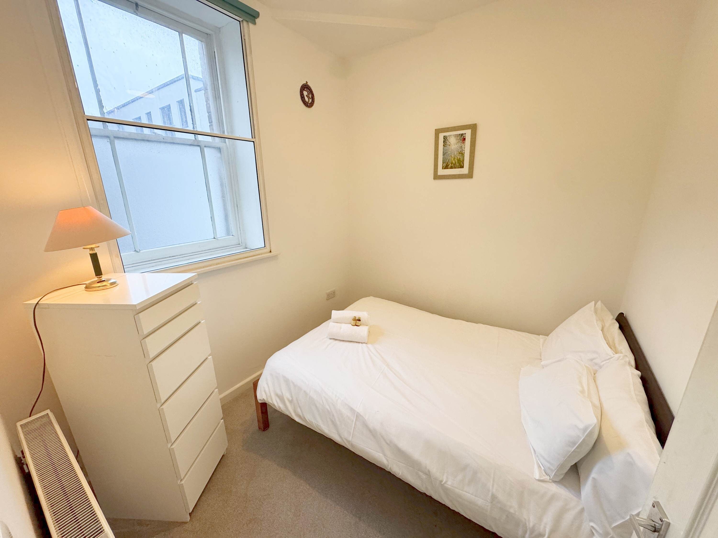 Een kleine, verzorgde slaapkamer met een enkel bed, nachtkastje met lamp, een kast en een radiator.