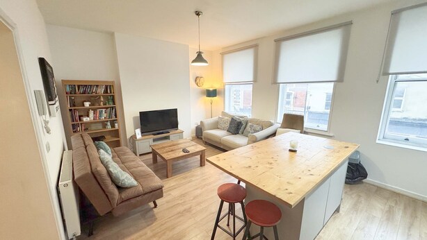 Een knus woongedeelte met een bank, een salontafel, een televisie, een boekenkast en een klein keukentje met krukjes.