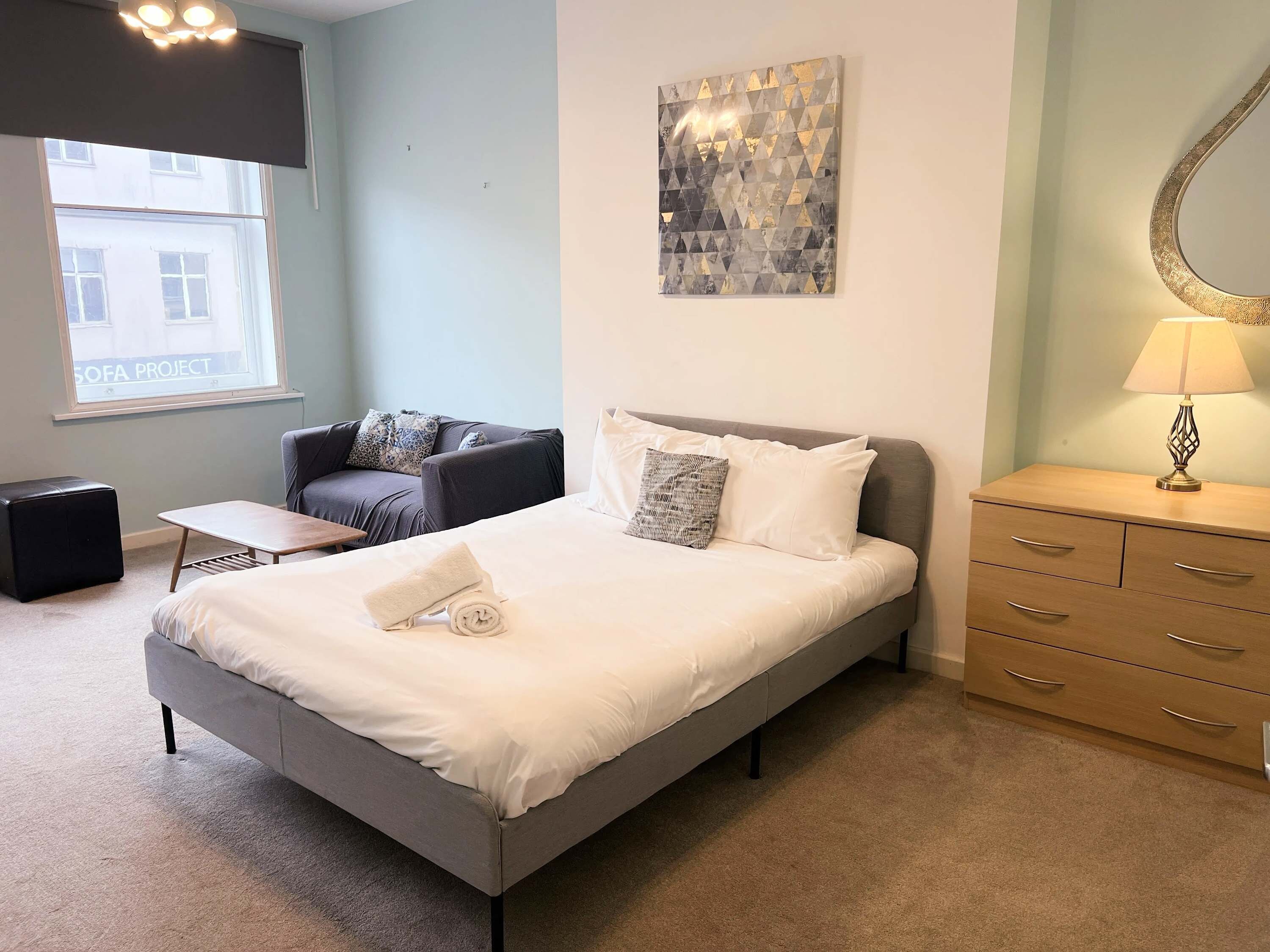 A bedroom with a bed, a sofa, a small table, a dresser, and a window with a view of buildings.