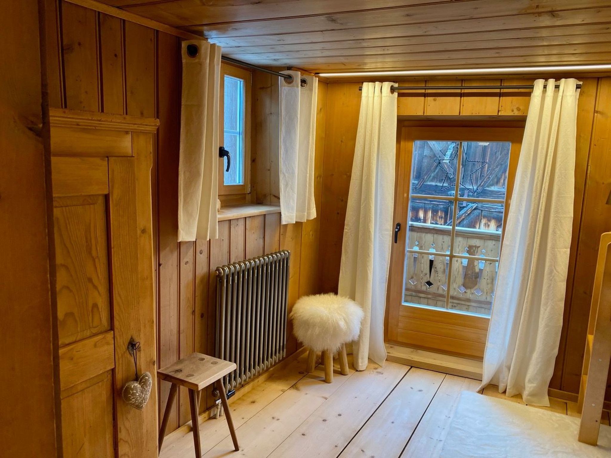 Interior em madeira com radiador, um banquinho e janela com cortinas.