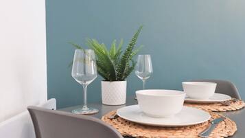 A dining table set with white plates, bowls, and cutlery, accompanied by two wine glasses and a potted fern.