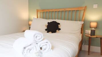A neatly made bed with rolled white towels, a black stuffed toy, and a bedside lamp on a wooden nightstand.