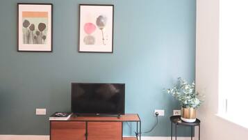 A room with a TV on a wooden stand, two framed artworks on the wall, a small table with a plant, and a power outlet.