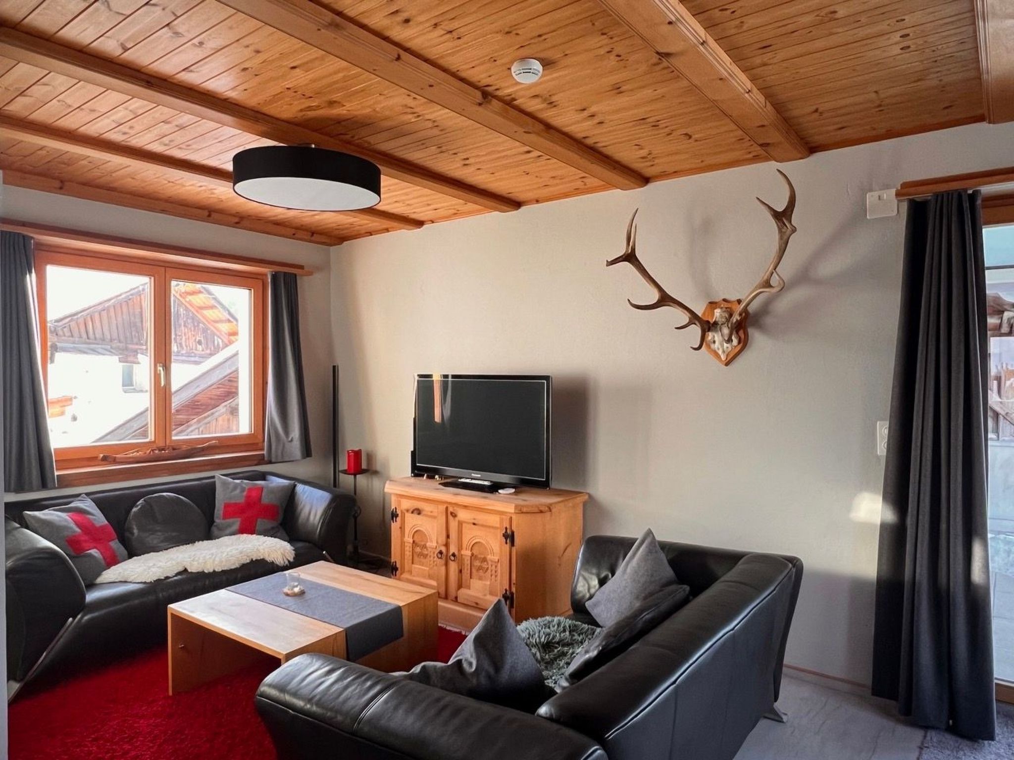 A cozy living room with a wooden ceiling, a mounted deer antler, a television on a wooden cabinet, and a red carpet.