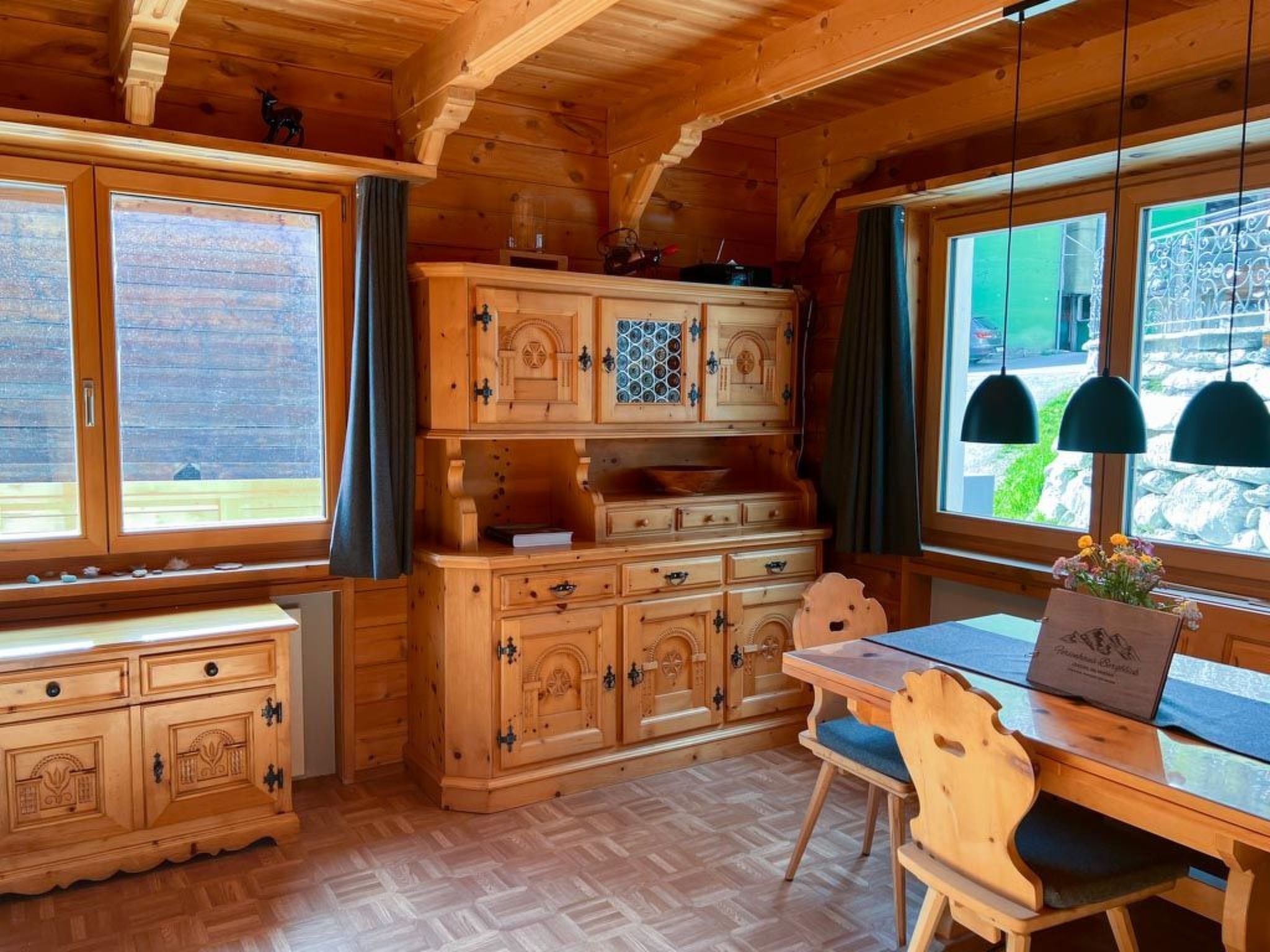 Ein Holzblockhaus-Interieur mit einem Esstisch, Stühlen, einem Schrank mit Ziergriffen und einem Fenster mit Vorhängen.