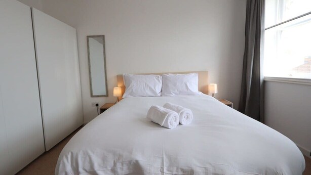A neatly made bed with white linens and rolled towels, a bedside table with a lamp, and a window with curtains. - Stylish 2 Bed in Old Market (Bristol)