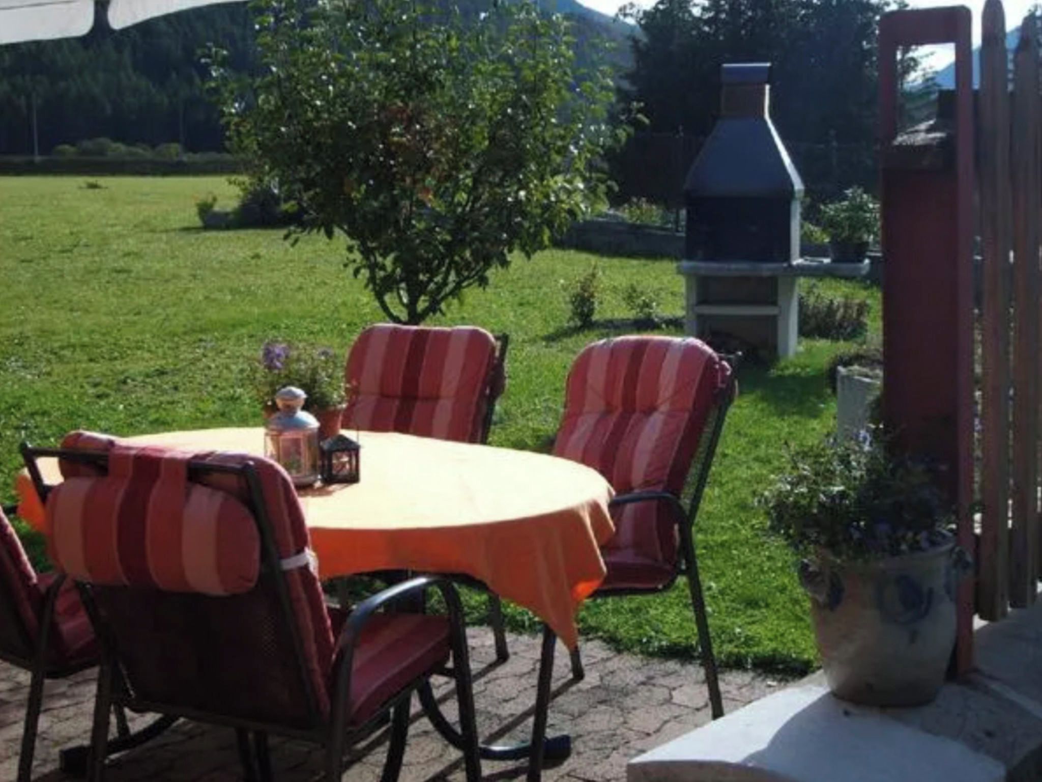 Eine Terrasse im Freien mit einem runden Tisch, gestreiften Stühlen, viel Grün und einem Grill.