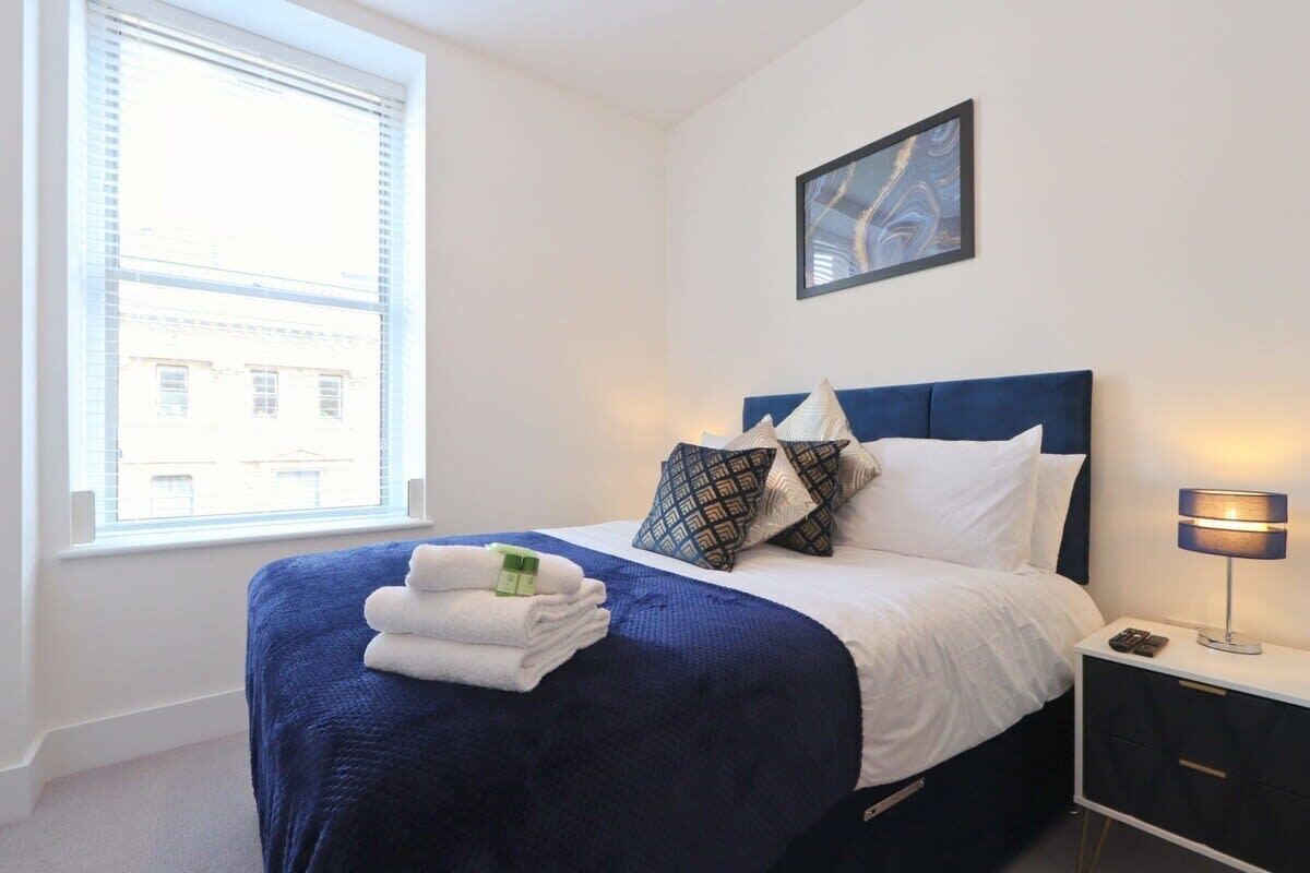 A neatly made bed with a blue headboard, white bedding, and a set of folded towels. A bedside table with a lamp and a remote control is visible.
