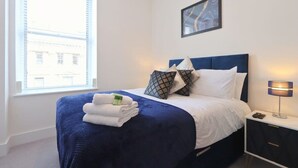 Un letto rifatto con cura, con una testiera blu, biancheria da letto bianca e un set di asciugamani piegati. Si nota un comodino con una lampada e un telecomando.