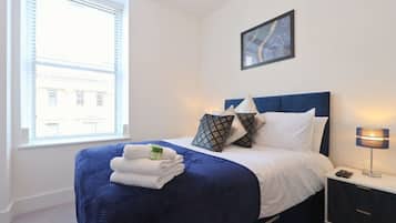 A neatly made bed with a blue headboard, white bedding, and a set of folded towels. A bedside table with a lamp and a remote control is visible.