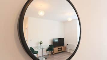 A room with a round mirror, a glass table, two green chairs, a TV on a wooden stand, and a small plant.