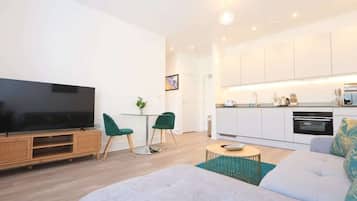 A modern living room with a flat-screen TV, green chairs, a sofa, and a kitchen area with white cabinets and a built-in oven.