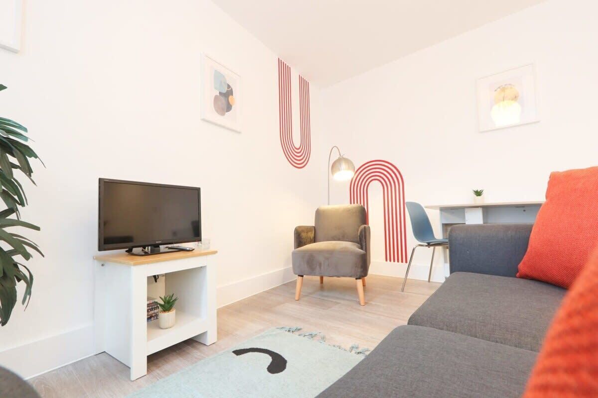 Un salon moderne avec un téléviseur à écran plat, un canapé gris, un coussin rouge décoratif et un bureau avec une chaise.