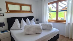 A bedroom with a bed, a nightstand, a lamp, a window with curtains, and a ceiling light fixture. - Scandella, (Sent) by Interhome (Sent)