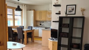 A modern kitchen with wooden cabinets, a dining area with a round table, and a black shelving unit. - Scandella, (Sent) by Interhome (Sent)