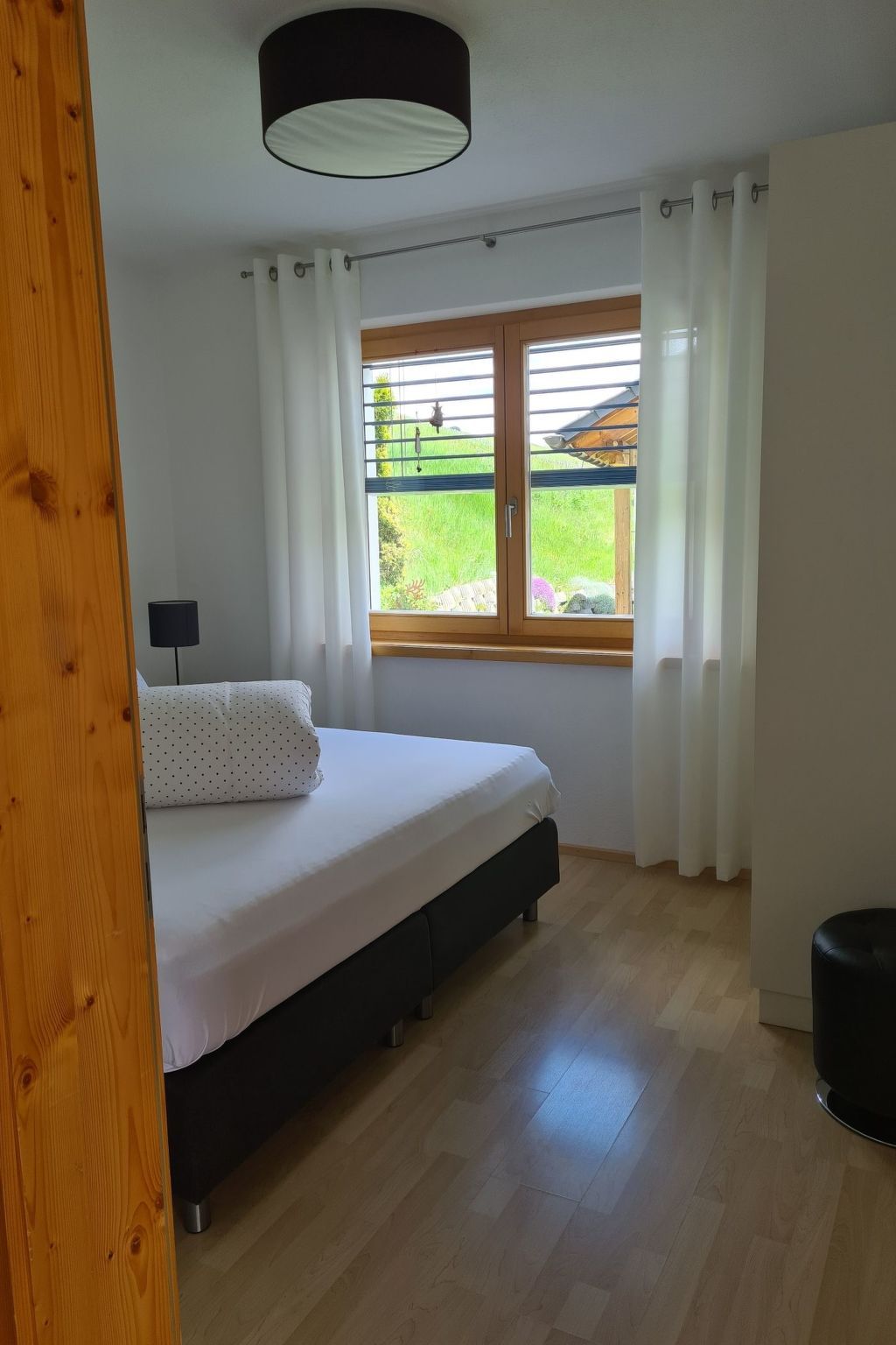 Ein Schlafzimmer mit einem Bett, einem Fenster mit Vorhängen, einer Lampe und Blick auf Grünfläche.