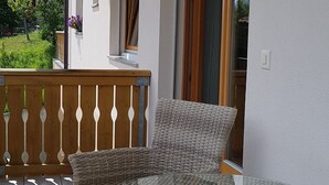 A wicker outdoor seating area with a glass table, a wooden fence, and a view of a building with a balcony. - Scandella, (Sent) by Interhome (Sent)