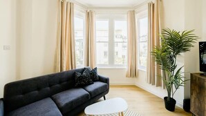 Una sala de estar con un sofá oscuro, una mesa de centro blanca, una planta en maceta y un televisor sobre un mueble de madera.