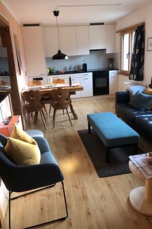 Ein modernes Wohnzimmer mit einem blauen Sofa, einem Holzes Esstisch und einer Küche im Hintergrund.