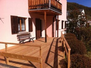 A wooden deck with a bench, a balcony with wooden railings, and a building with a pink facade.