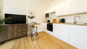 Una cocina moderna con armarios blancos, encimera de madera, microondas, estufa y fregadero. Hay un taburete y un televisor sobre un mueble de madera.