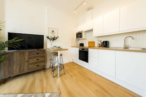 A modern kitchen with white cabinets, a wooden countertop, a microwave, a stove, and a sink. There is a bar stool and a television on a wooden cabinet. - Grange Rd Flat 2 one bedroom flat (Bristol)