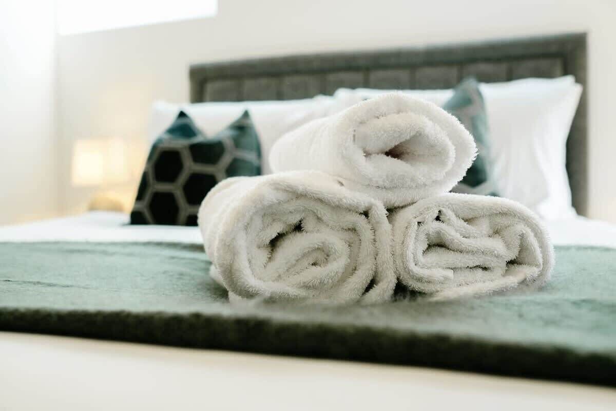 A neatly made bed with rolled white towels and a green blanket, a patterned pillow, and a headboard.