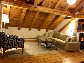 Uma sala de estar aconchegante no estilo loft, com teto inclinado, vigas de madeira, um sofá, uma poltrona estampada e uma mesa de centro de vidro.