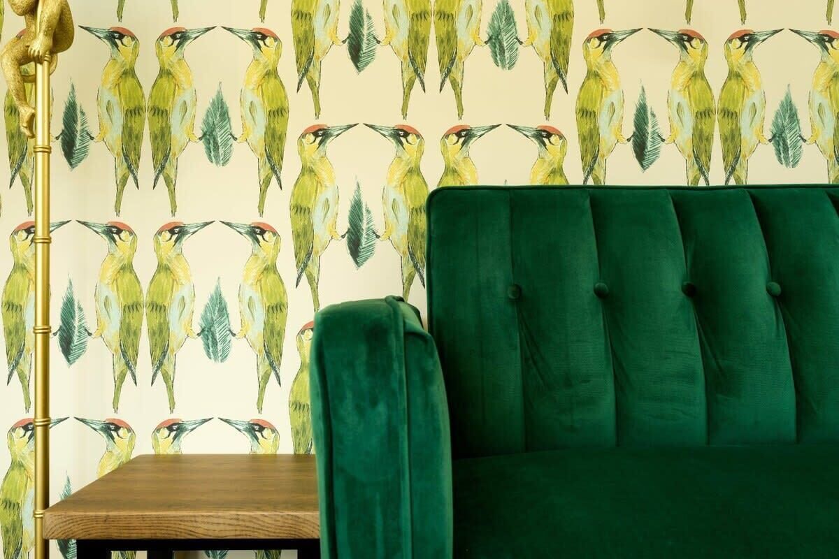 Une pièce avec un canapé recouvert d’un tissu vert, une table en bois et un papier peint orné d’un motif répétitif représentant des oiseaux.