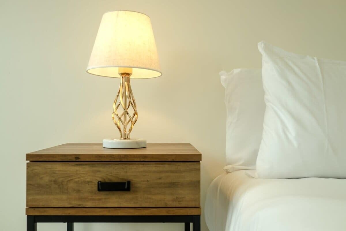 Een houten nachtkastje met een lamp, een lade en een wit kussen op een bed.
