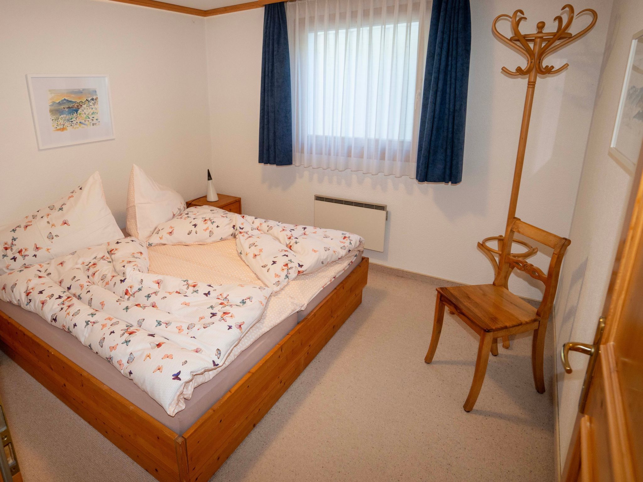 Een slaapkamer met een bed, nachtkastje, stoel, radiator en een raam met gordijnen.