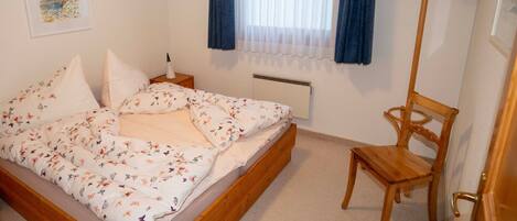 Een slaapkamer met een bed, nachtkastje, stoel, radiator en een raam met gordijnen.