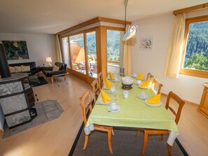 Uma área de refeições com mesa posta para quatro pessoas, cadeiras de madeira e vista para uma paisagem montanhosa através de amplas janelas.