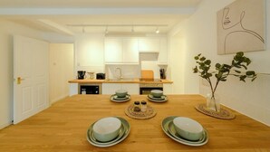 A modern kitchen with white cabinets, a wooden dining table set with plates, and a vase with a plant on the right. - Grange Road Two Bedroom Flat (Bristol)