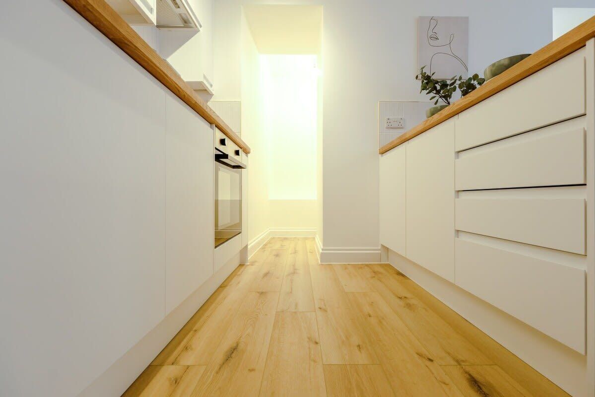 Cocina estrecha con suelo de madera, armarios blancos y techo de madera.