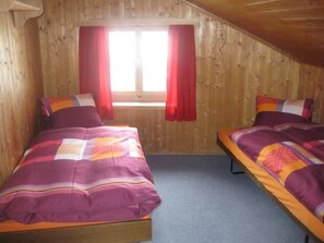 A room with two beds, wooden walls, a window with red curtains, and a sloped ceiling. - Chasa Munt Fallun 3, (Ftan) by Interhome (Ftan)
