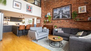 A spacious living area with a kitchen, grey sofas, a coffee table, and a large black and white cityscape painting on the brick wall. - The Robinson Building (Bristol City)