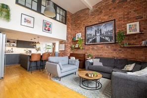A spacious living area with a kitchen, grey sofas, a coffee table, and a large black and white cityscape painting on the brick wall. - The Robinson Building (Bristol City)