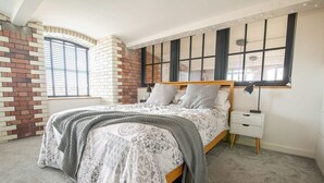 A bedroom with a bed, a nightstand, a lamp, a window with blinds, and a brick wall. - The Robinson Building (Bristol City)