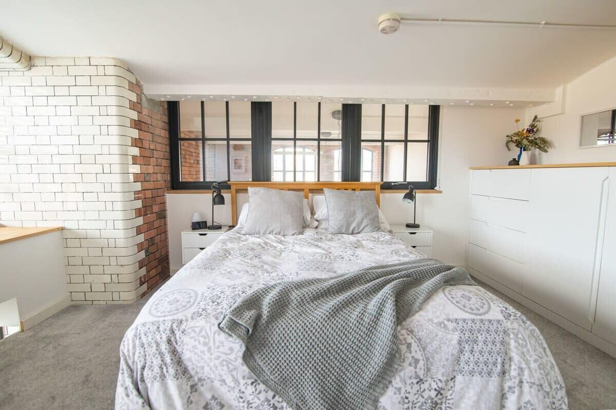 A bedroom with a brick wall, a bed with a patterned bedspread, a nightstand, a dresser, and a window with blinds.