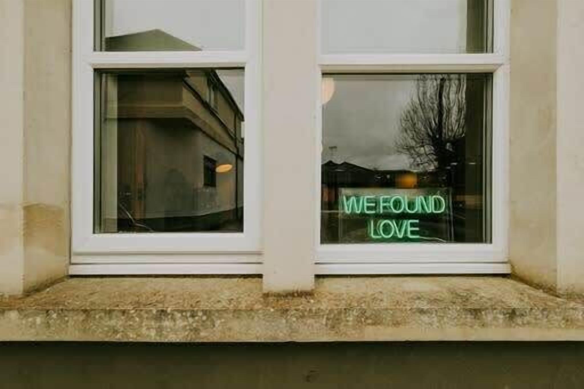 A building with two windows, one of which has a neon sign that reads 'WE FOUND LOVE'.